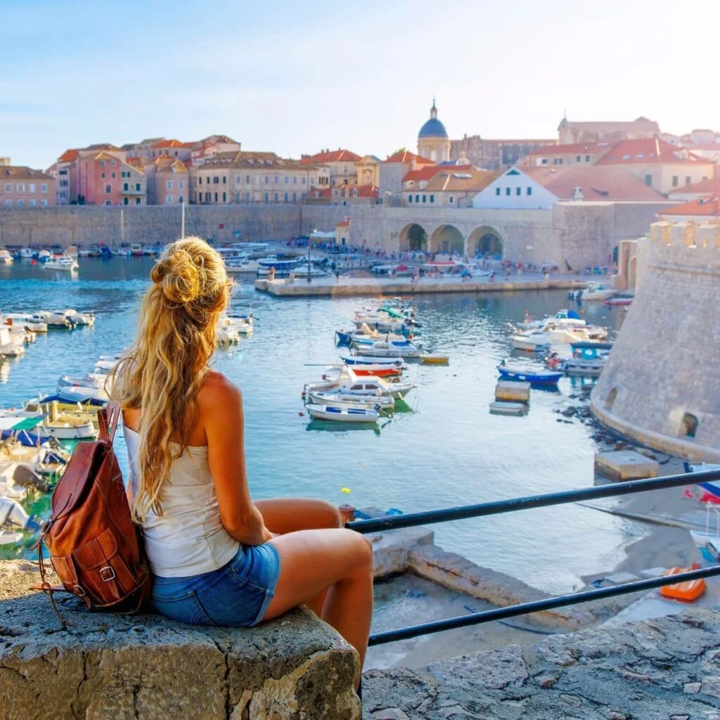 A Female Tourist Sat On The Ramparts Of Dubrovnik Admiring A View Of The Old Town, Croatia