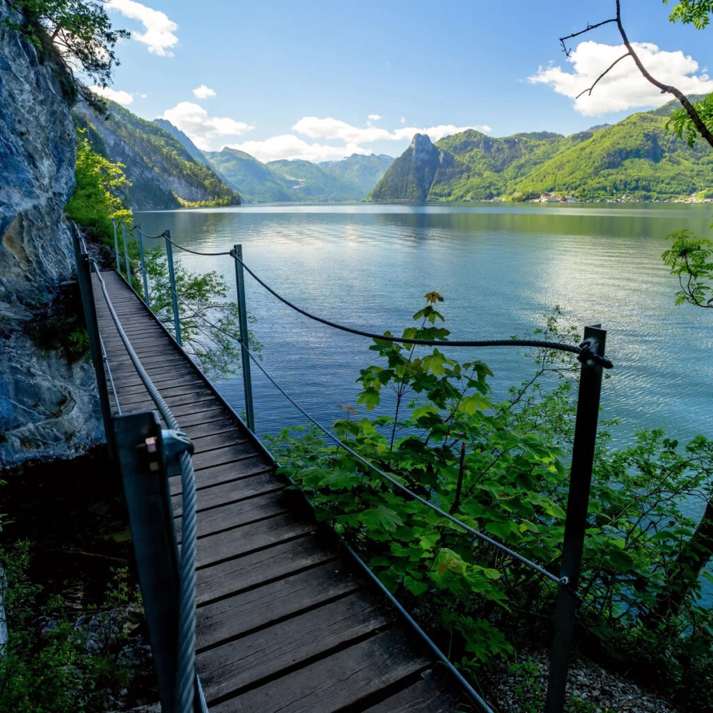 Wooden footbridge on the Miesweg hiking trail above Lake Traunsee, surrounded by lush spring vegetation and dramatic limestone mountains in Austria's Salzkammergut region.