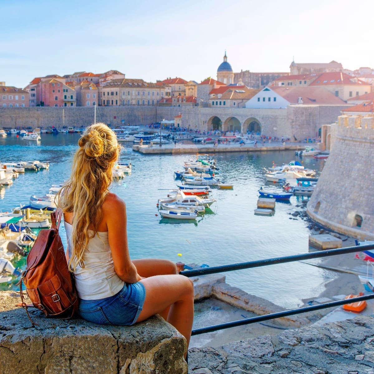A Female Tourist Sat On The Ramparts Of Dubrovnik Admiring A View Of The Old Town, Croatia